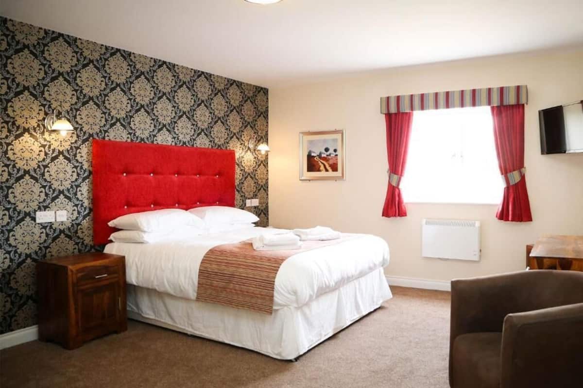 Une chambre avec un grand lit, une table de chevet, une chaise, une fenêtre avec des rideaux, un téléviseur fixé au mur et un papier peint à motifs.