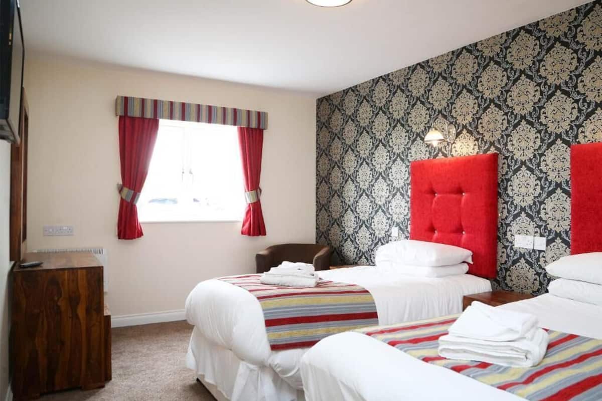 Une chambre d’hôtel avec deux lits, une tête de lit rouge, un papier peint à motifs et une fenêtre avec des rideaux rouges.