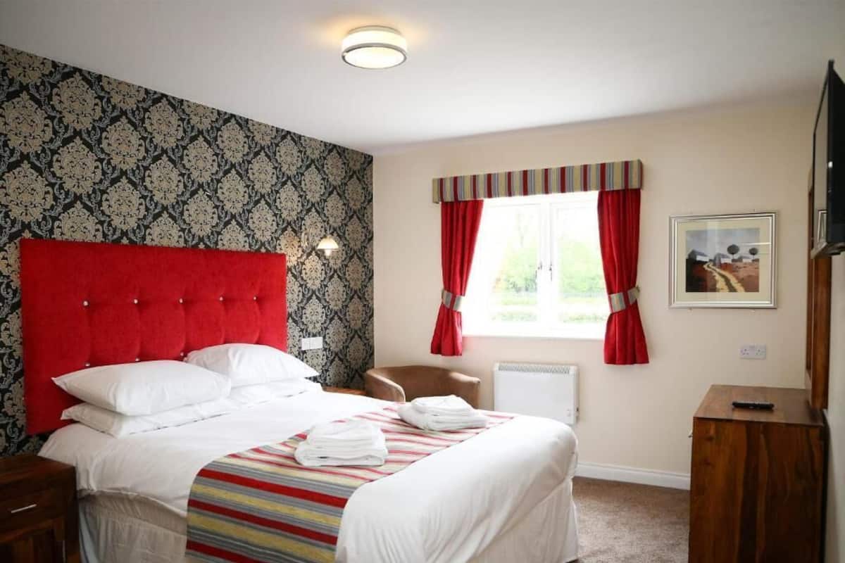 Un lit bien fait, avec du linge de lit blanc, une tête de lit rouge et une couverture à rayures. Il y a une fenêtre avec des rideaux rouges, une chaise et un petit meuble en bois.