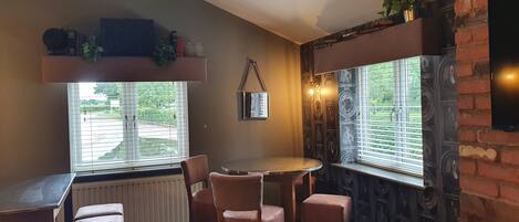 Una habitación acogedora con una mesa redonda y cuatro sillas, un radiador, una ventana con persianas y una pared de ladrillo con detalles decorativos.