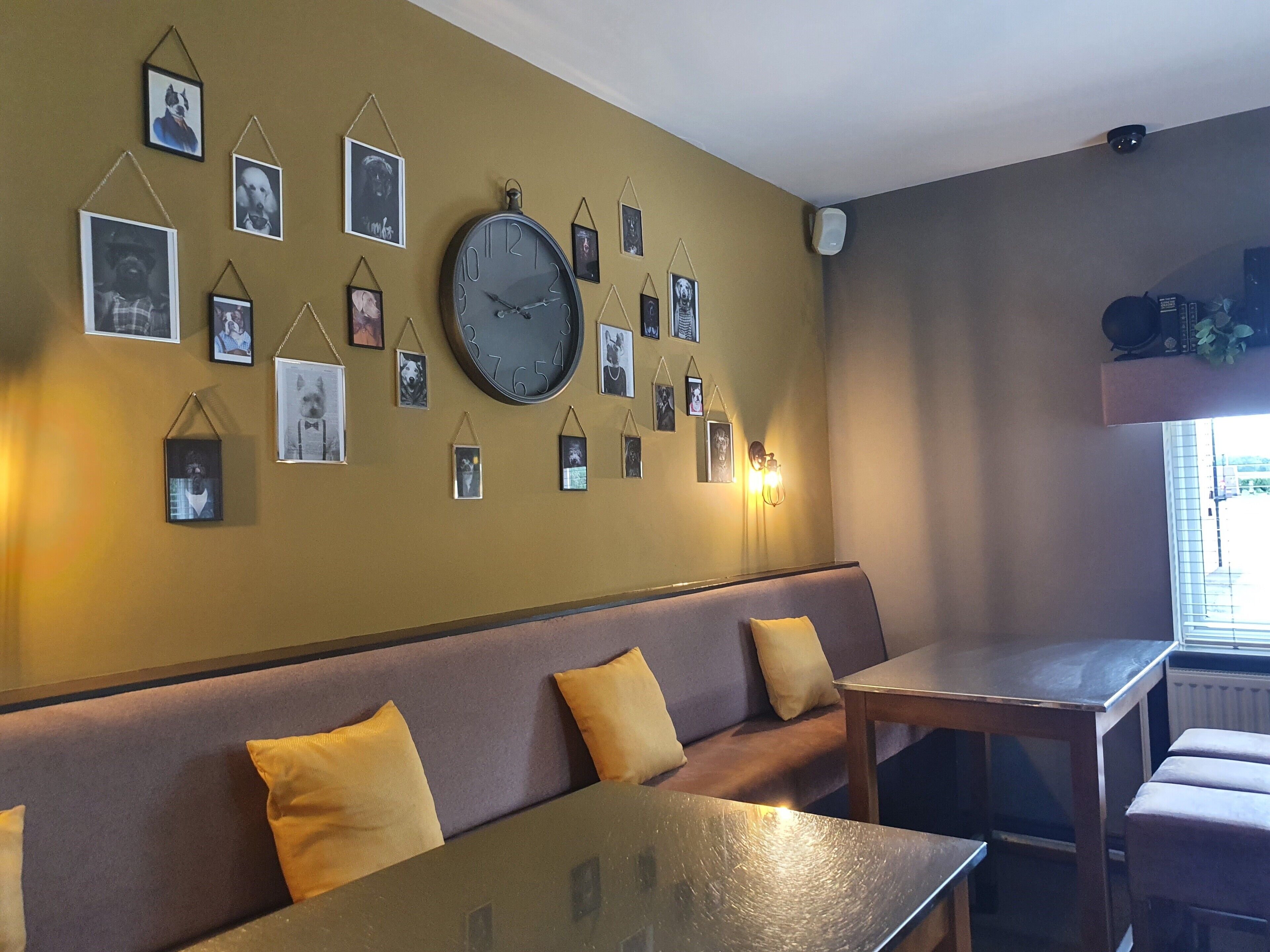 A room with a large wall clock, multiple framed pictures, a sofa with yellow cushions, and a table.