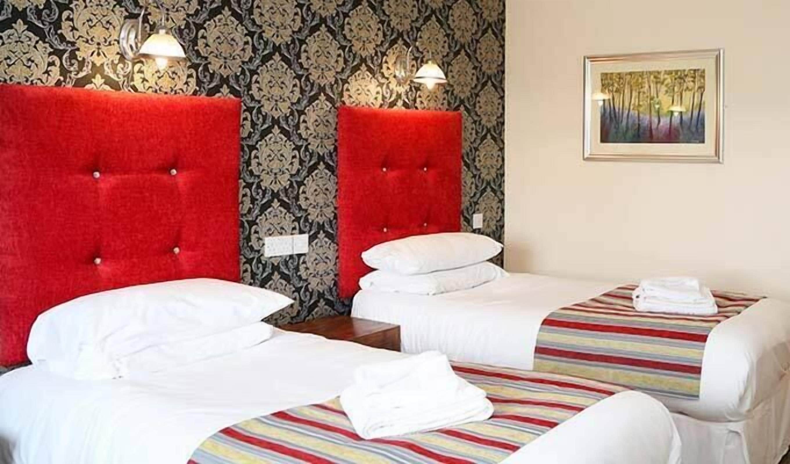 A hotel room with two beds, each with a red headboard, white bedding, and striped bedspreads. There is a framed picture on the wall and a lamp on the nightstand.