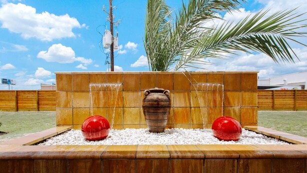 A water feature with two red spheres and a central urn-like structure, surrounded by a tiled wall and palm trees. - Palace Inn and Suites - Willowbrook Mall (Houston)