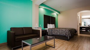 A hotel room with a brown sofa, a bed with a patterned bedspread, a small table, and a green accent wall. - Palace Inn and Suites - Willowbrook Mall (Houston)