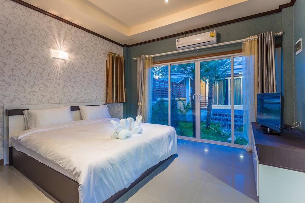 A modern bedroom with a large bed, a flat-screen TV, and a sliding glass door leading to a garden.