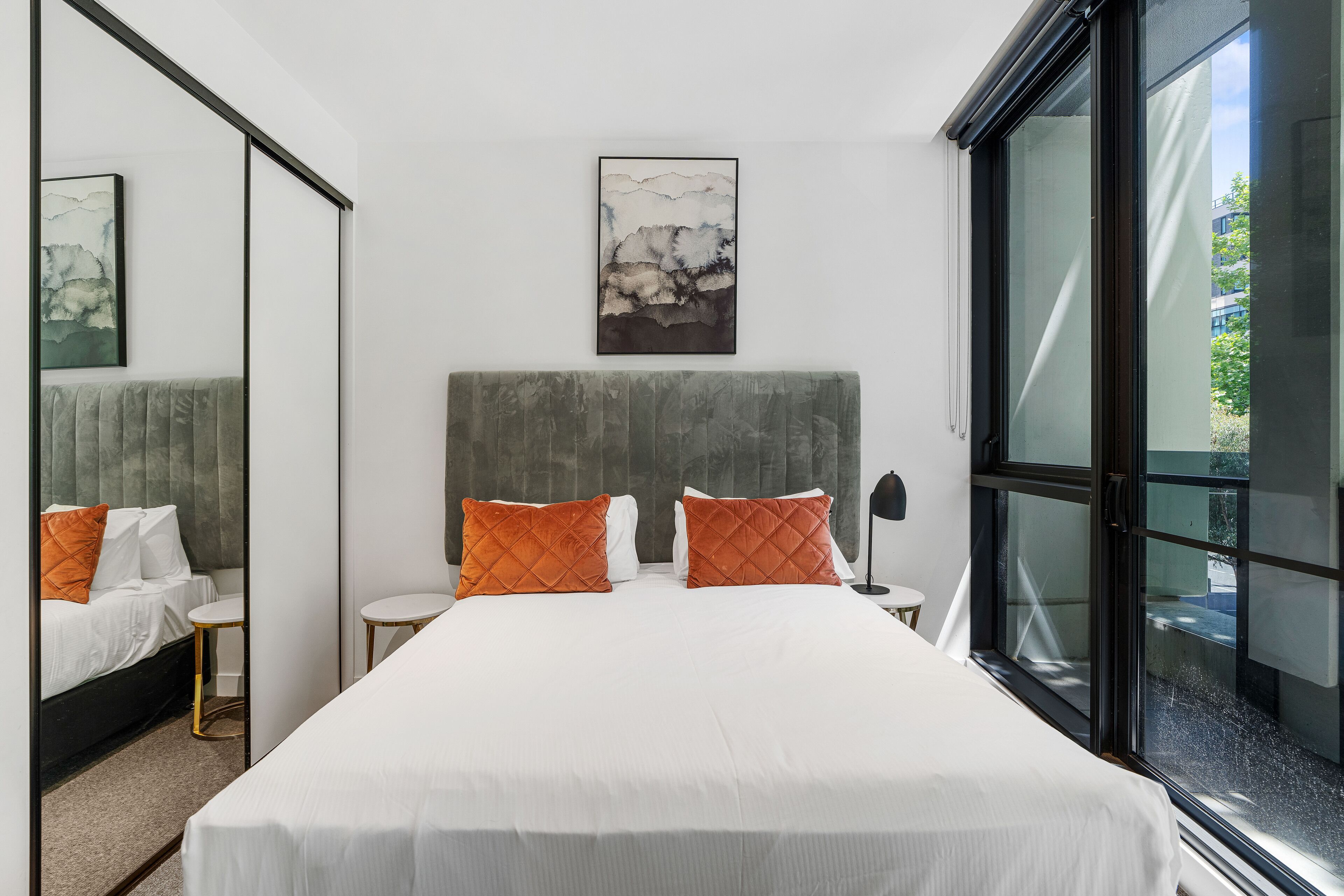 A modern hotel room with a large bed, two orange pillows, a gray headboard, a white wall, a framed artwork, and a large window.