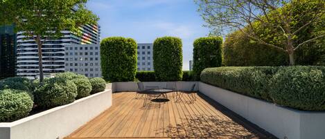 Una terraza en la azotea con piso de madera, una mesa redonda con sillas y setos bien cuidados.