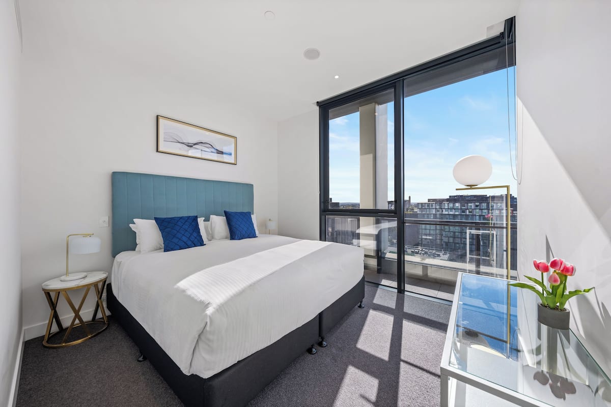 Una habitación de hotel moderna con una cama grande, una mesita auxiliar pequeña, una mesa de vidrio con planta y vistas a la ciudad a través de amplias ventanas.