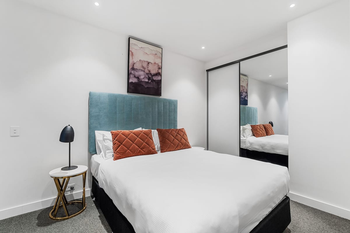 Una habitación de hotel moderna con una cama grande, una mesita redonda, un espejo en la pared y un cuadro sobre la cama.