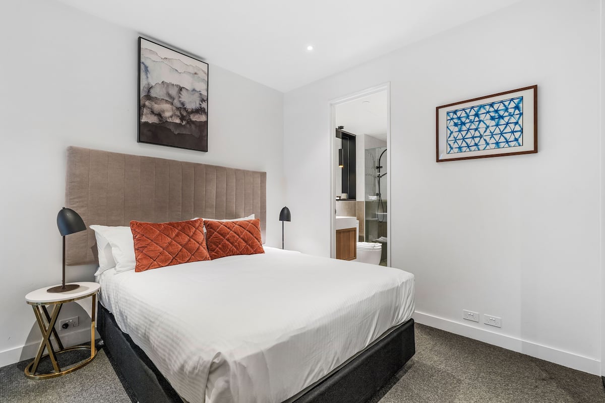 Un dormitorio moderno con una cama grande, una mesita de noche, una lámpara de pared y dos obras de arte enmarcadas.