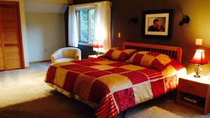 A bedroom with a large bed, a chair, a nightstand, a lamp, a window with curtains, and a framed picture on the wall.