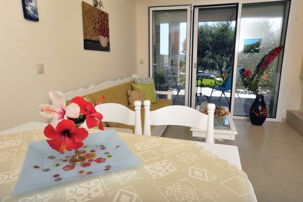 Un coin repas avec une table dressée pour deux personnes, une chaise blanche et un canapé avec des coussins. Un vase rempli de fleurs est posé sur la table.