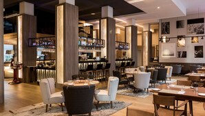 Un restaurante moderno con techos altos, vigas a la vista y una zona de bar con estanterías llenas de botellas. Las mesas están dispuestas con manteles blancos y sillas.