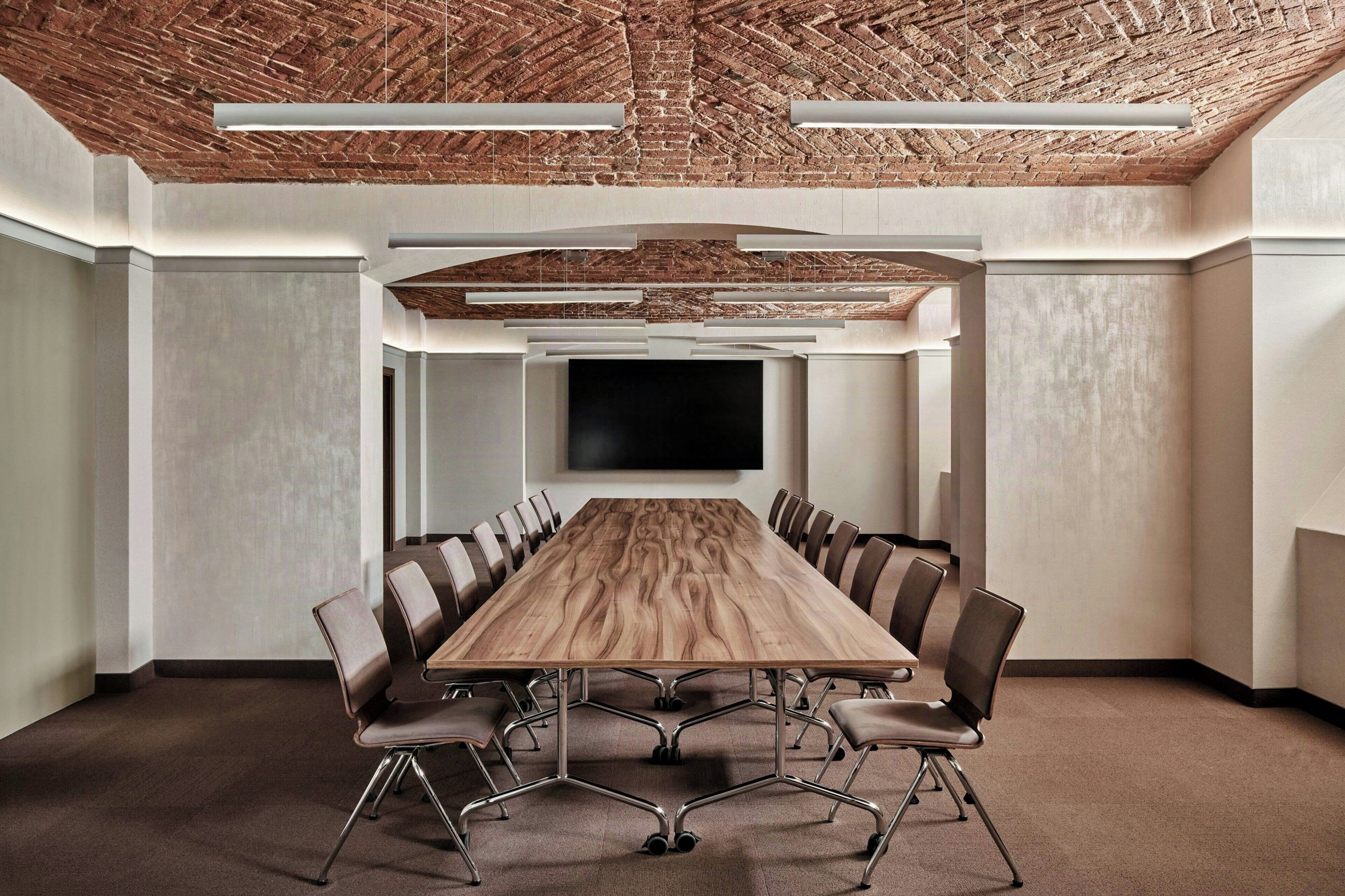 A conference room with a long table, chairs, a flat-screen TV, and a brick ceiling.