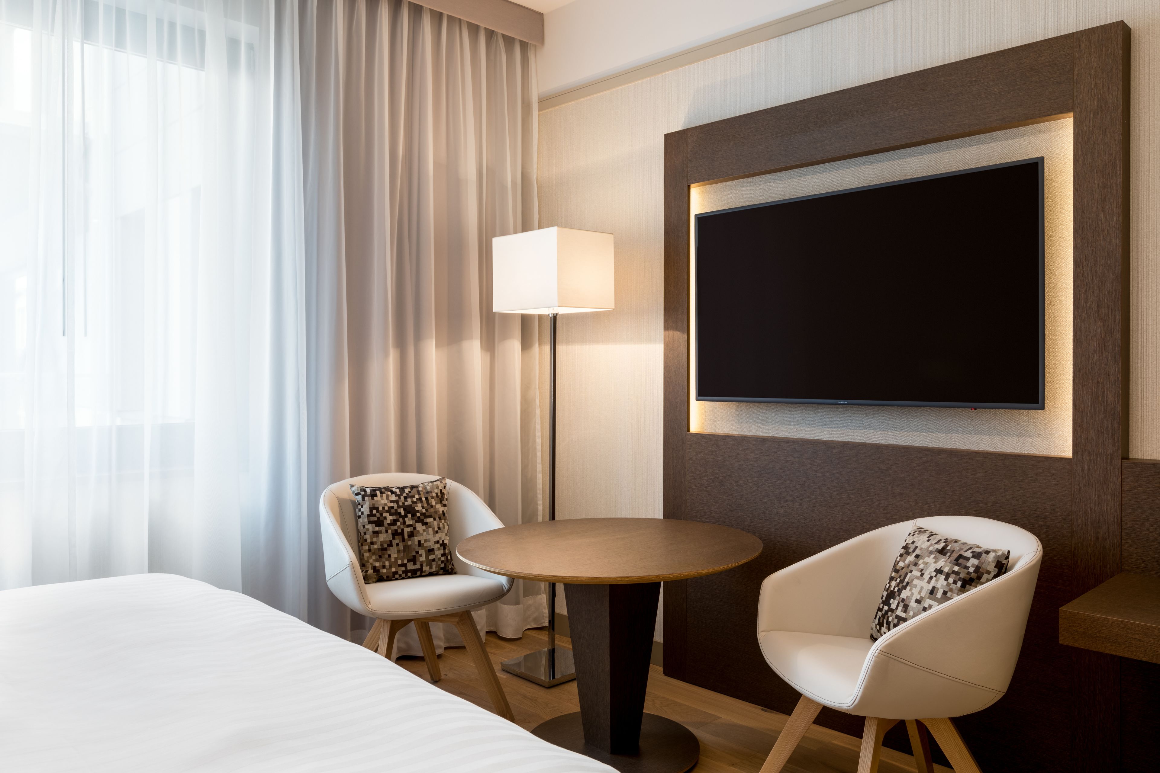 A hotel room with a bed, two chairs, a table, a TV, and a lamp.