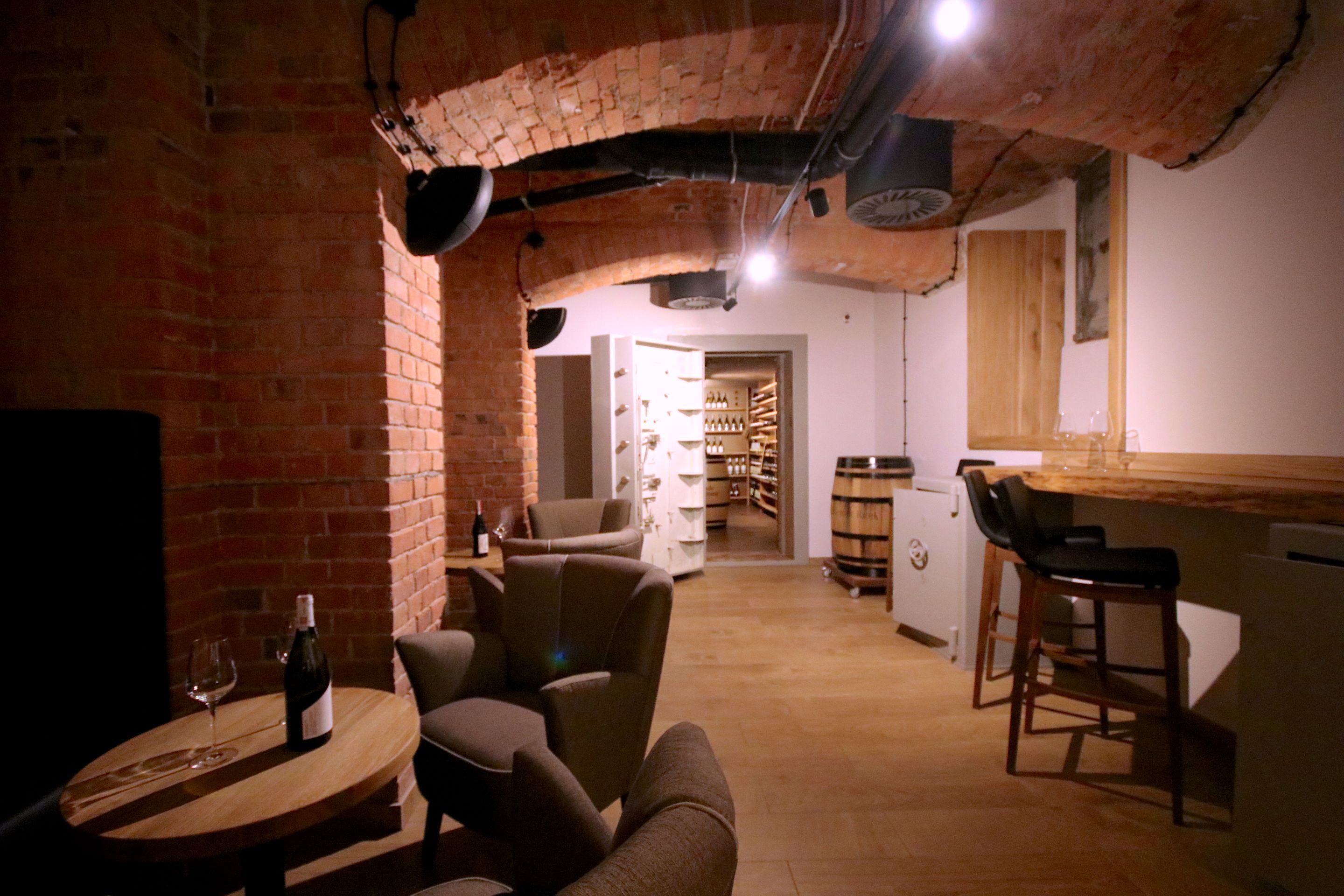 A cozy, dimly lit room with brick walls, a wooden table with a bottle of wine and glasses, and a bar area with stools.