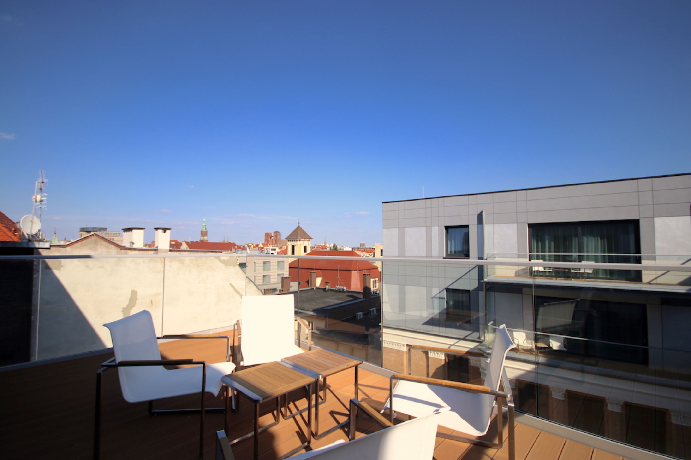 Une terrasse sur le toit, avec une table en bois et des chaises blanches, offrant une vue sur un paysage urbain dominé par des bâtiments modernes.