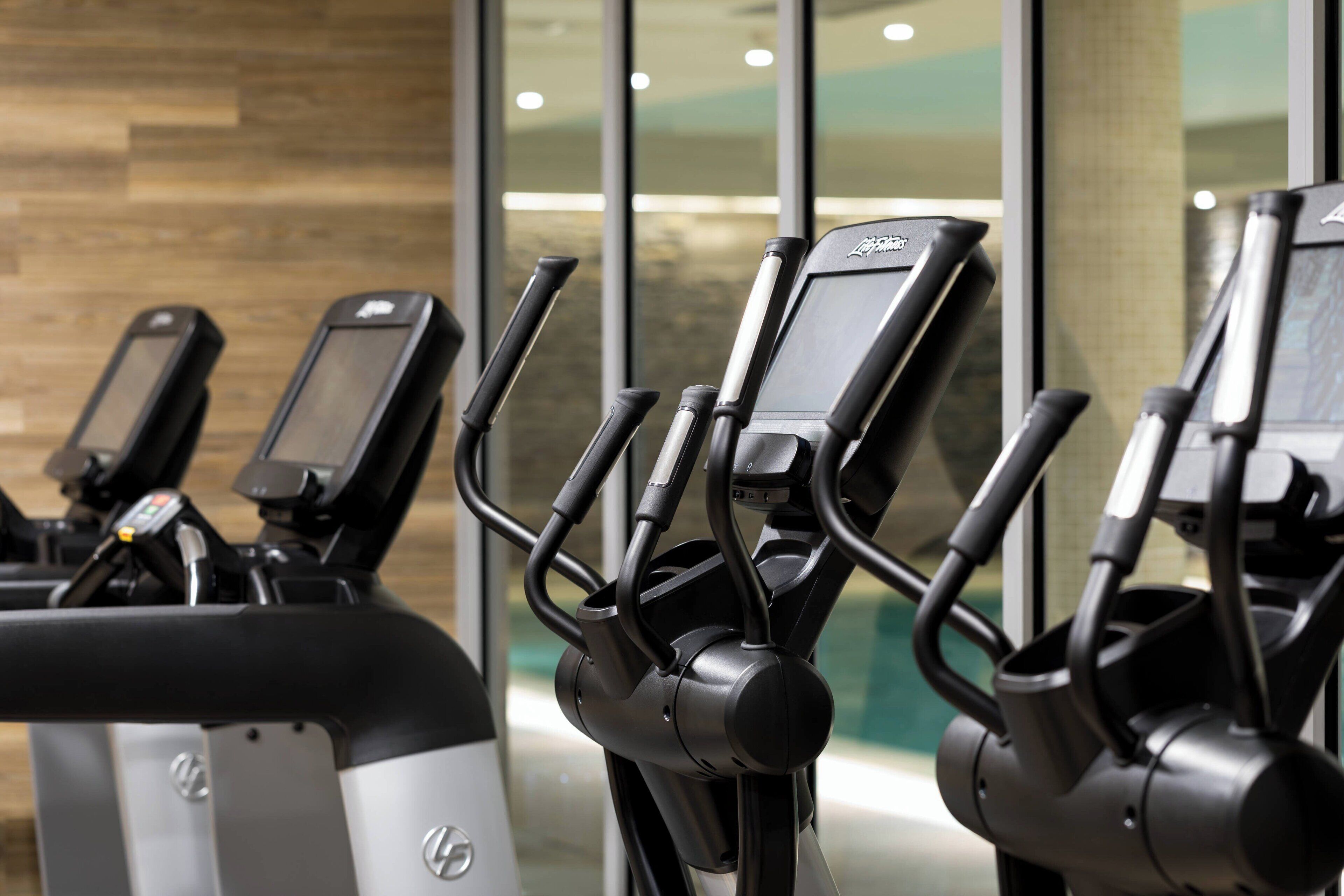 Une rangée de vélos d’appartement équipés d’écrans numériques, disposés dans une salle de sport dotée d’un plancher en bois et de grandes fenêtres.