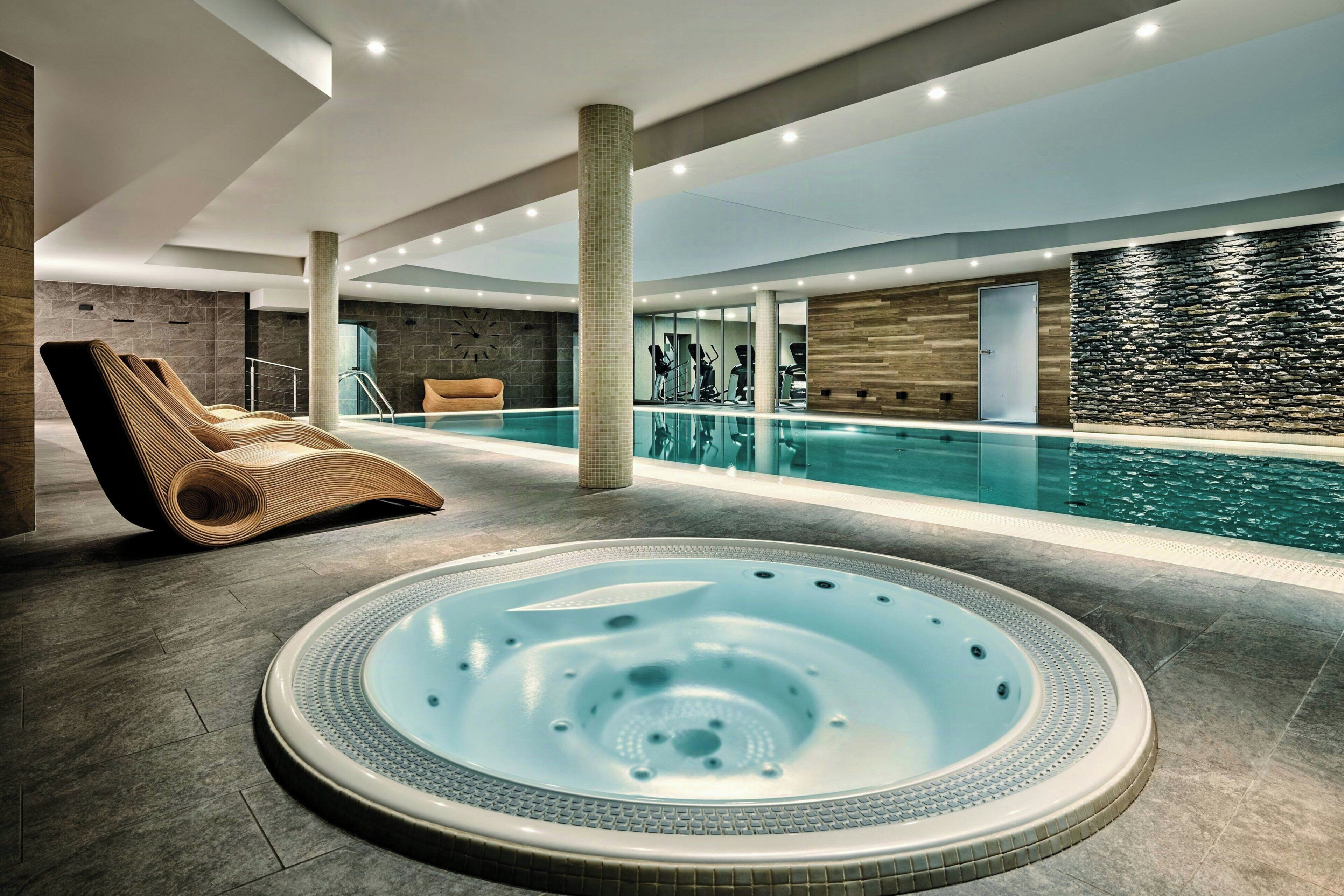 A modern indoor swimming pool area with a hot tub, exercise equipment, and a lounge chair.