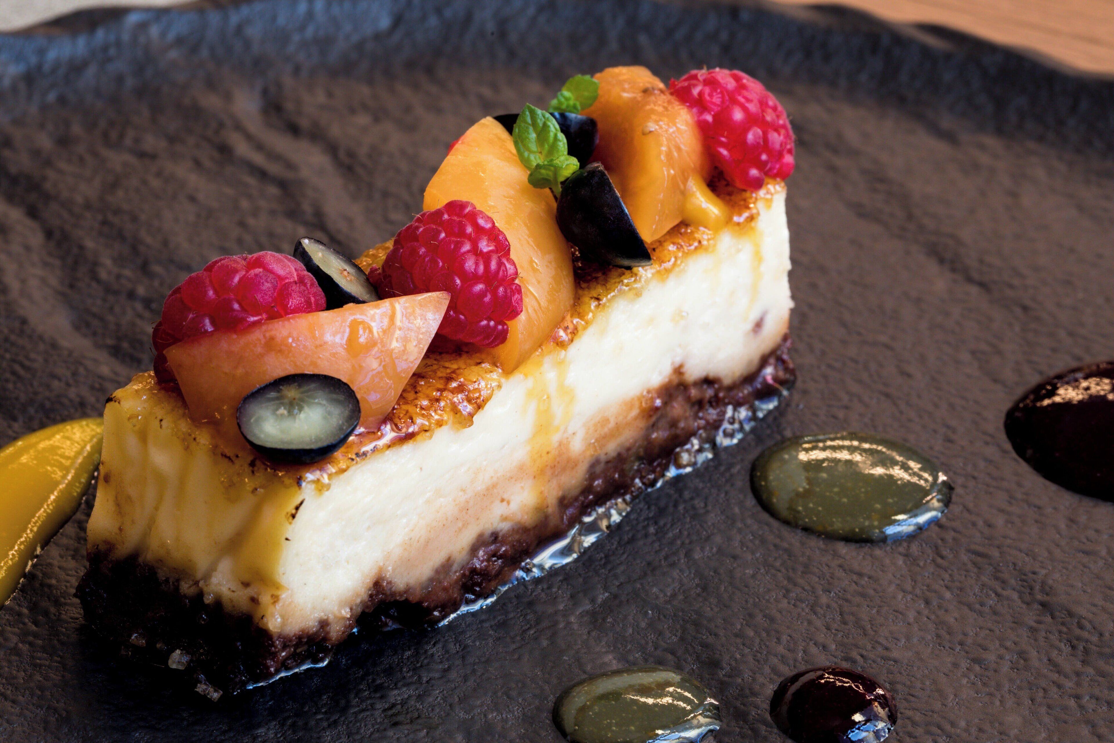 A slice of cheesecake topped with fruits and a mint leaf on a dark slate plate.