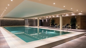 Una piscina interior moderna con un fondo de pared de piedra, iluminación empotrada y sillones de descanso.