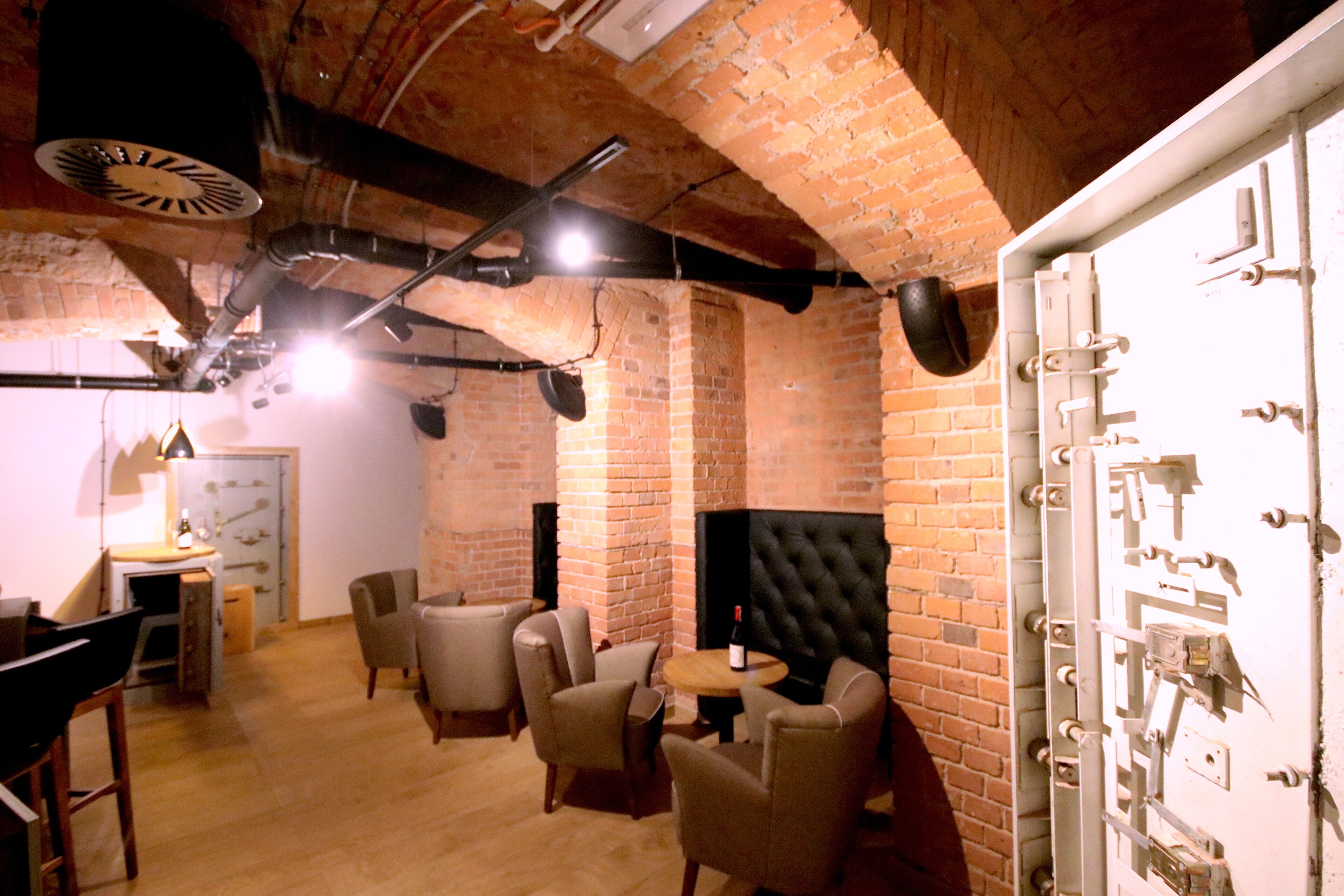 A room with brick walls, a fireplace, and a white door with multiple locks. There are several chairs and a small table with a bottle.