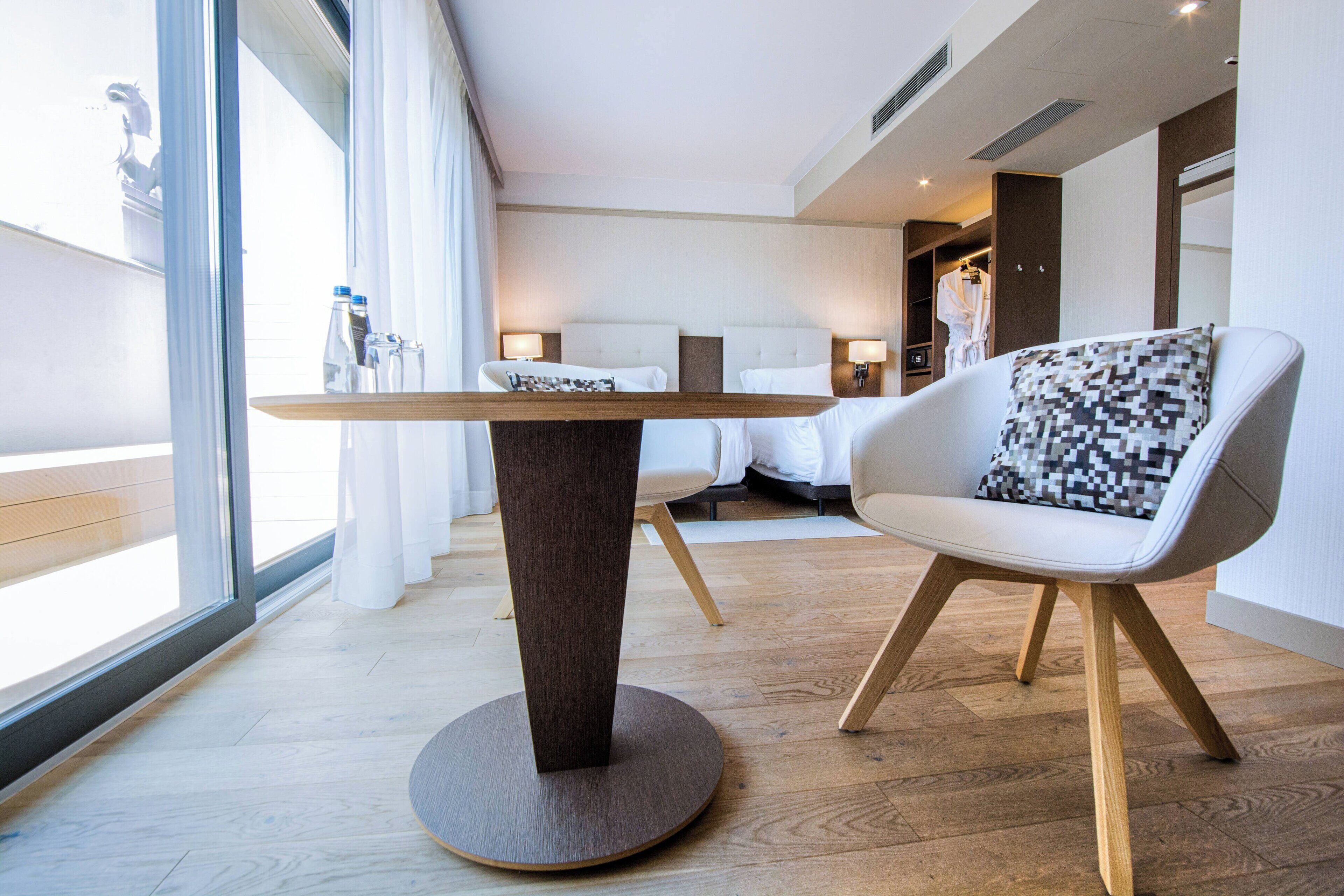 A modern hotel room with a round wooden table, a white armchair with a pixelated pillow, and a large window with curtains.