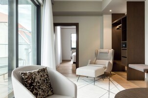 A modern hotel room with a large window, a white armchair, a sofa, a small table, and a built-in wardrobe.