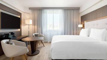 Habitación de hotel con una cama grande, un televisor de pantalla plana, un escritorio con una silla, una mesa redonda y una ventana con cortinas.