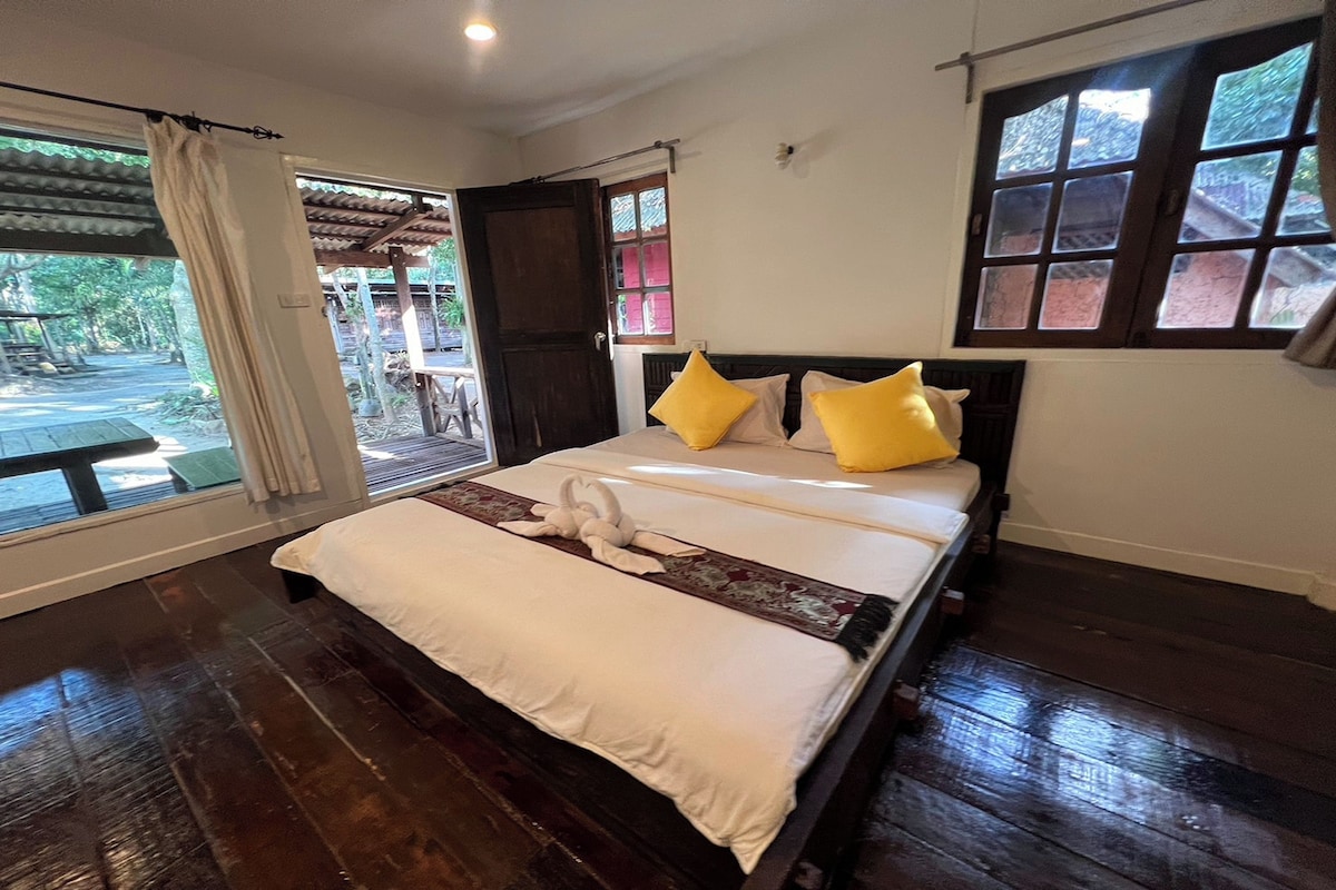 Una cama con sábanas blancas y dos almohadas amarillas, suelo de madera y una ventana con vistas al exterior.