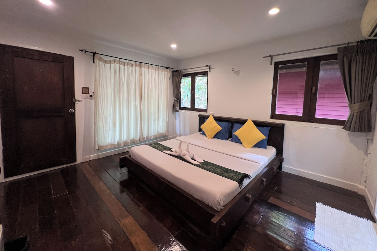 A bedroom with a wooden bed, white curtains, and a window with a view of greenery.