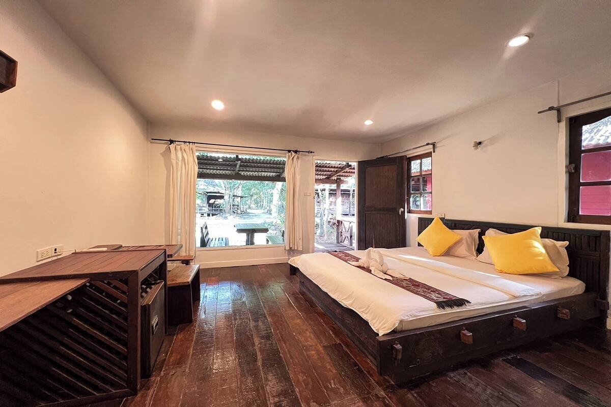 A bedroom with a wooden bed, a small desk, a window with curtains, and a view of the outside.