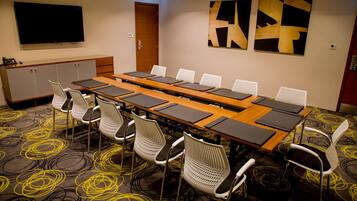 Un salón de conferencias con una larga mesa, sillas blancas, un televisor, arte abstracto en la pared y una alfombra estampada.