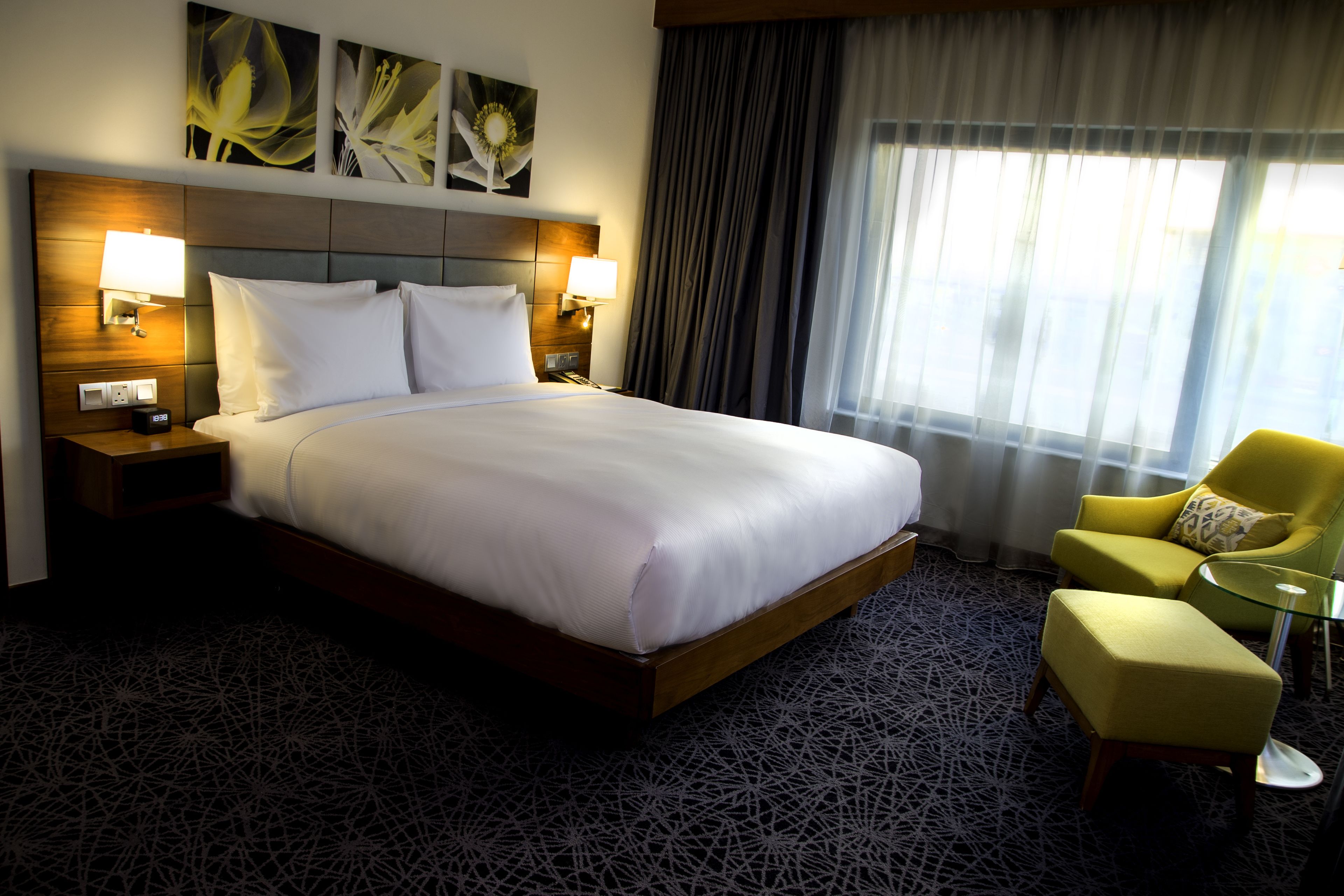 Quarto de hotel com uma cama grande, duas luminárias de cabeceira, um criado-mudo, uma poltrona amarela, uma mesa de vidro e uma janela com cortinas.