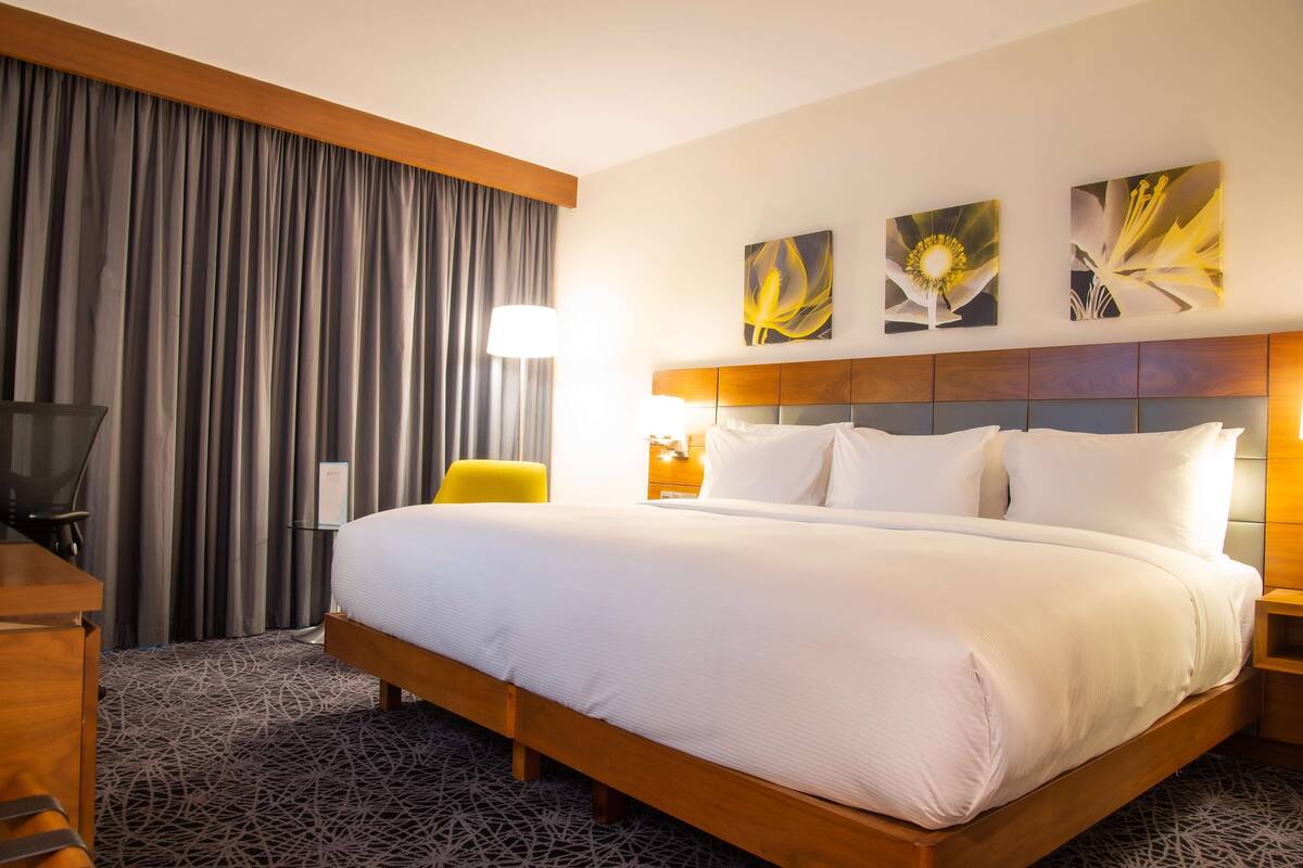 A hotel room with a large bed, a desk, a chair, and a TV. There are three paintings on the wall and a curtain on the window.