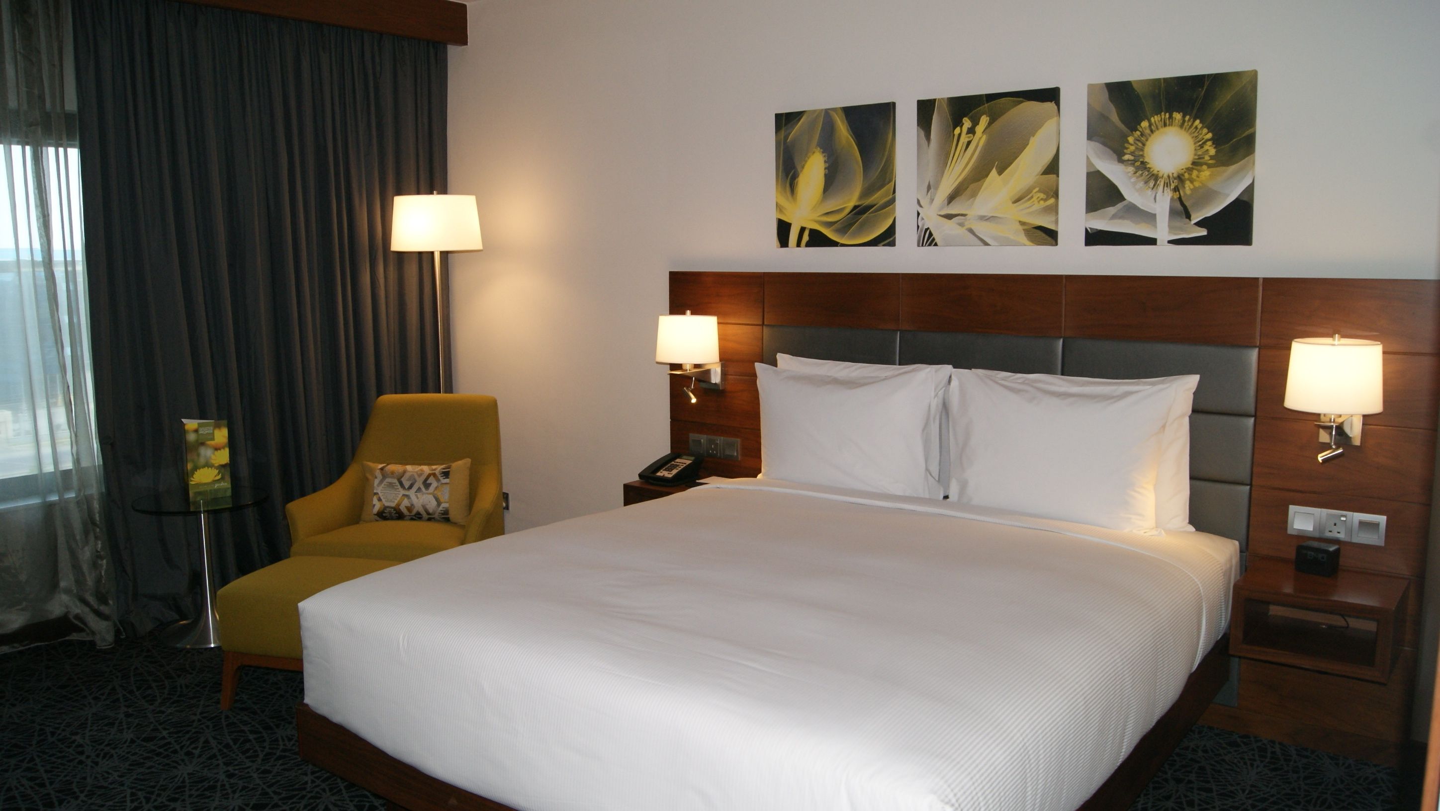 Habitación de hotel con una cama grande, dos lámparas de noche, una silla con cojín, una mesita de noche con teléfono y tres cuadros de flores colgados en la pared.
