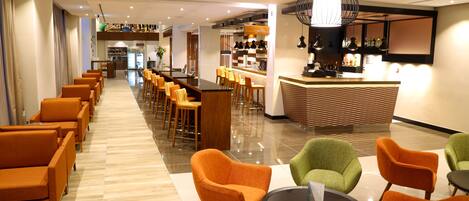 A hotel lobby with a long row of orange chairs, a reception desk, and a bar area.
