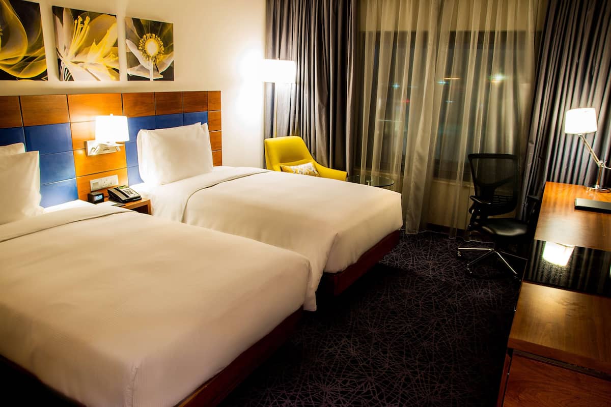 A hotel room with two beds, a desk, a chair, and a lamp.