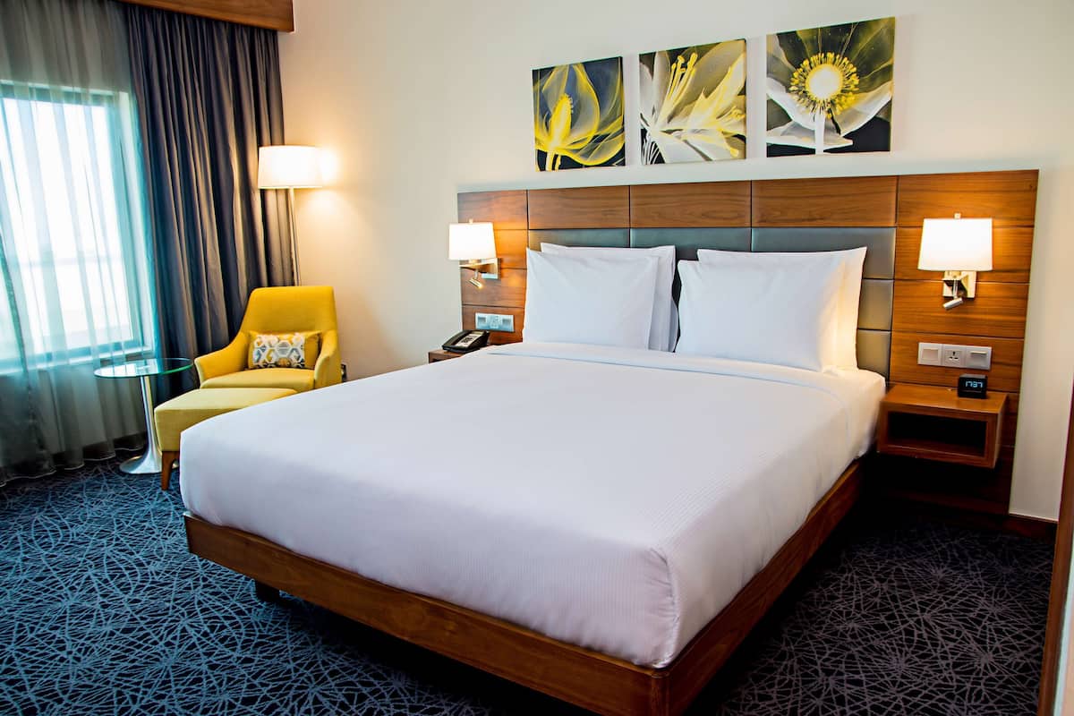 A hotel room with a large bed, two bedside tables with lamps, a small round table, a yellow armchair, and a window with curtains.
