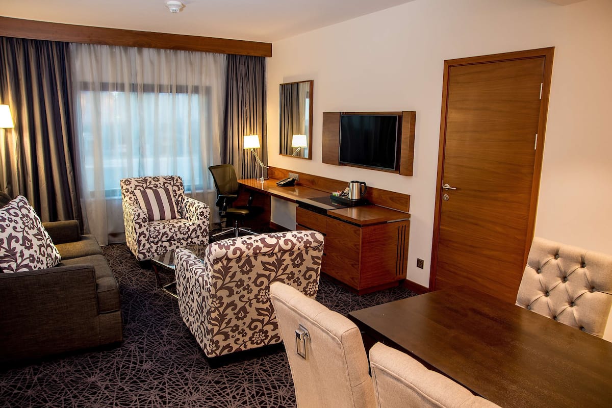 A hotel room with a sofa, armchair, and a dining table. There is a TV mounted on the wall and a door to the right.
