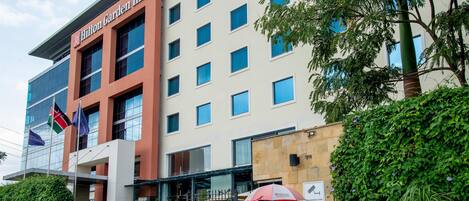A Hilton Garden Inn with a modern facade, multiple windows, and a well-maintained entrance area with greenery and a security booth.