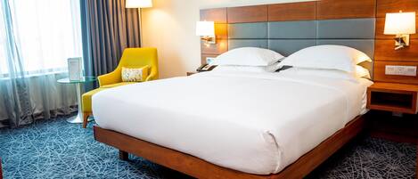 Un cuarto de hotel con una cama grande, un sillón amarillo, una mesita de vidrio y vistas al exterior a través de una ventana con cortinas.