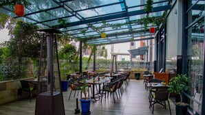 Un espacio cubierto para comer al aire libre con mesas y sillas, rodeado de plantas en macetas y una estructura de metal para la parrilla.