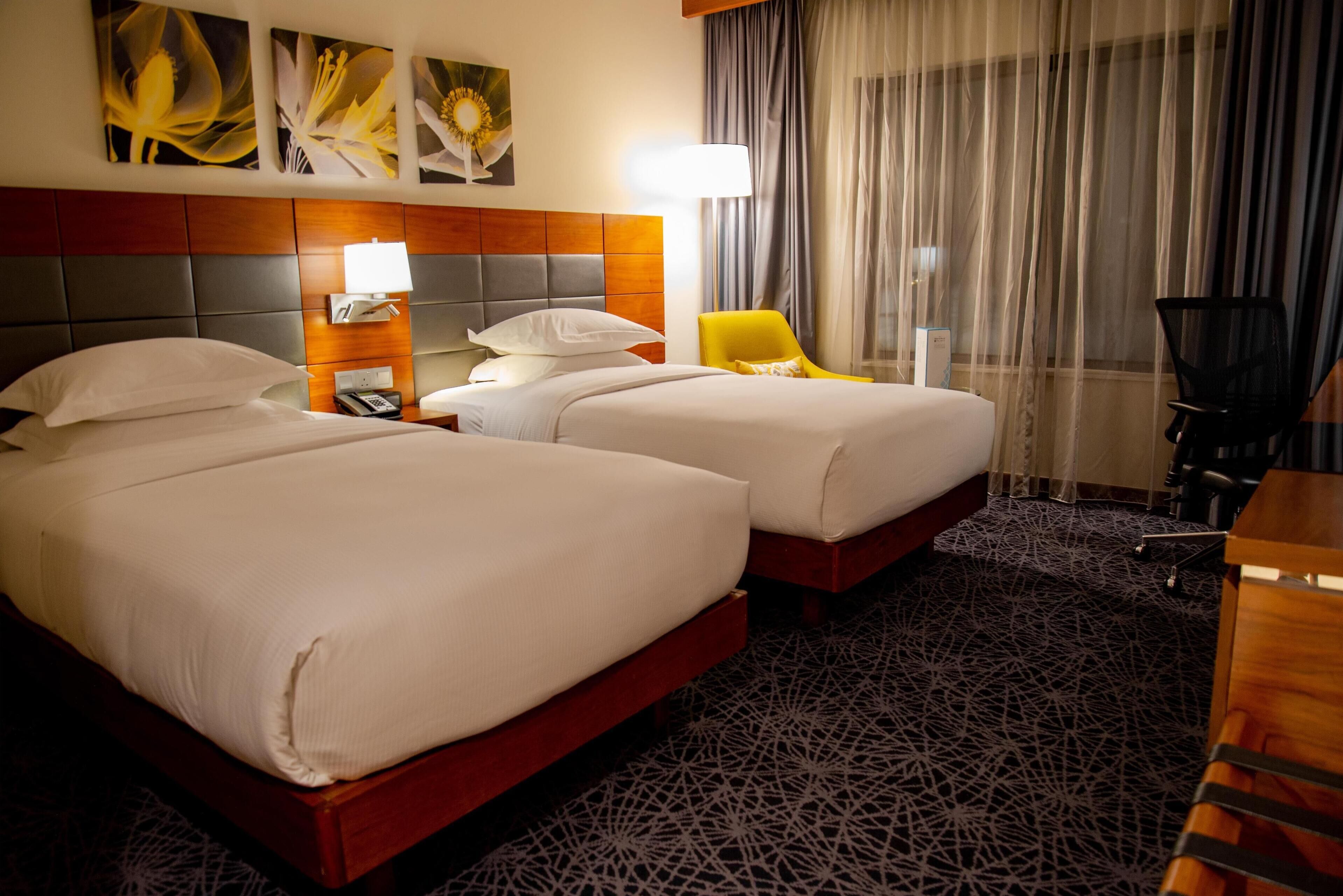 A hotel room with two beds, a wooden headboard, a patterned carpet, a yellow chair, and a desk with a lamp.
