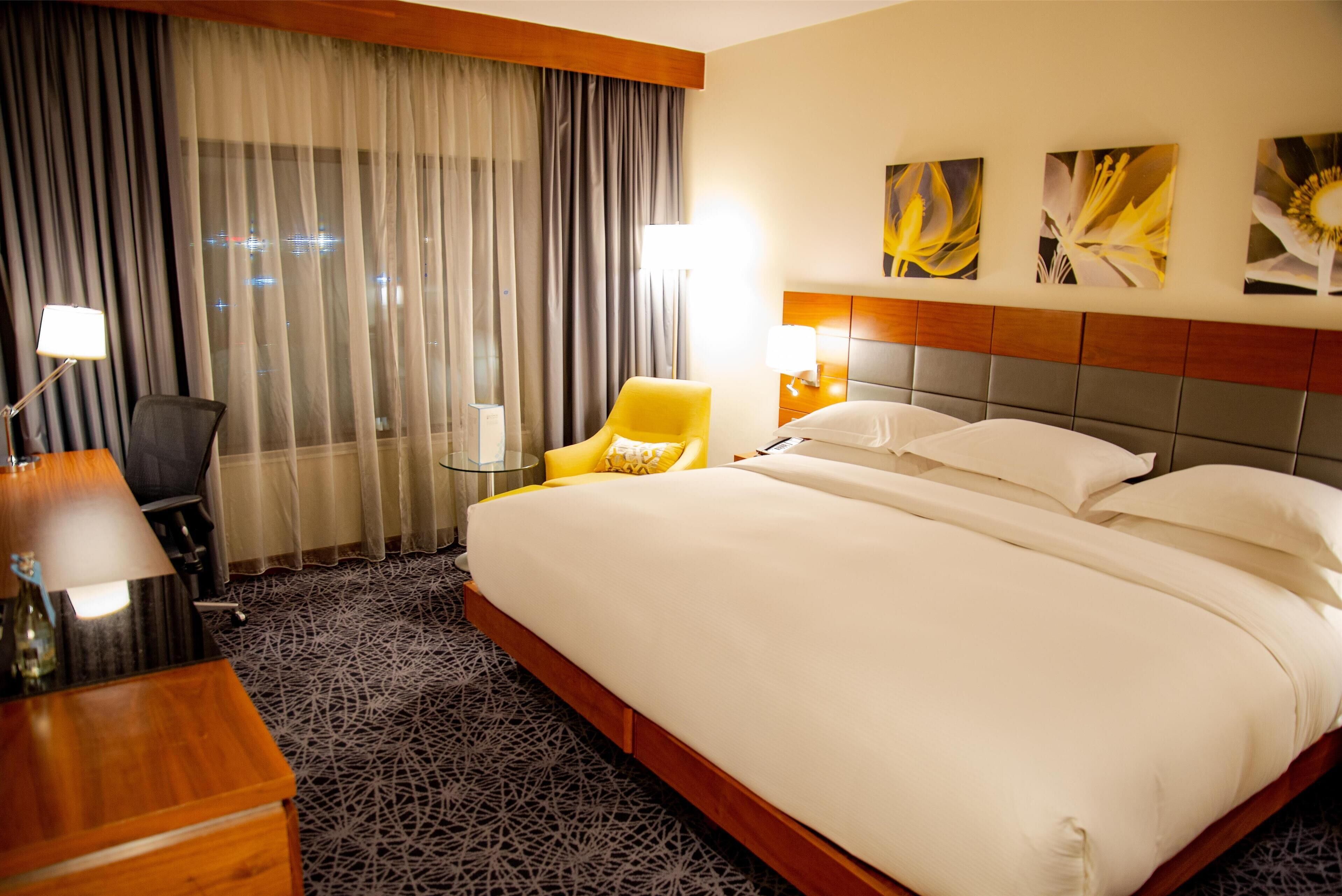 A hotel room with a large bed, a desk with a lamp, a chair, and a view of the city through sheer curtains.
