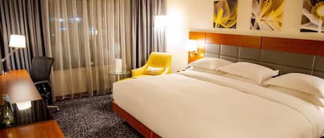 A hotel room with a large bed, a desk with a lamp, a chair, and a view of the city through sheer curtains.