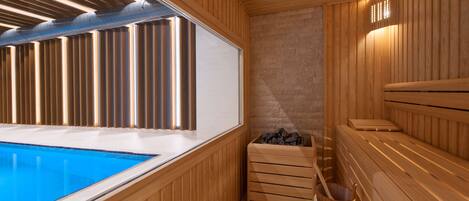 Una sauna con paredes de madera, un banco de madera, un balde pequeño de madera y una pared de piedra.