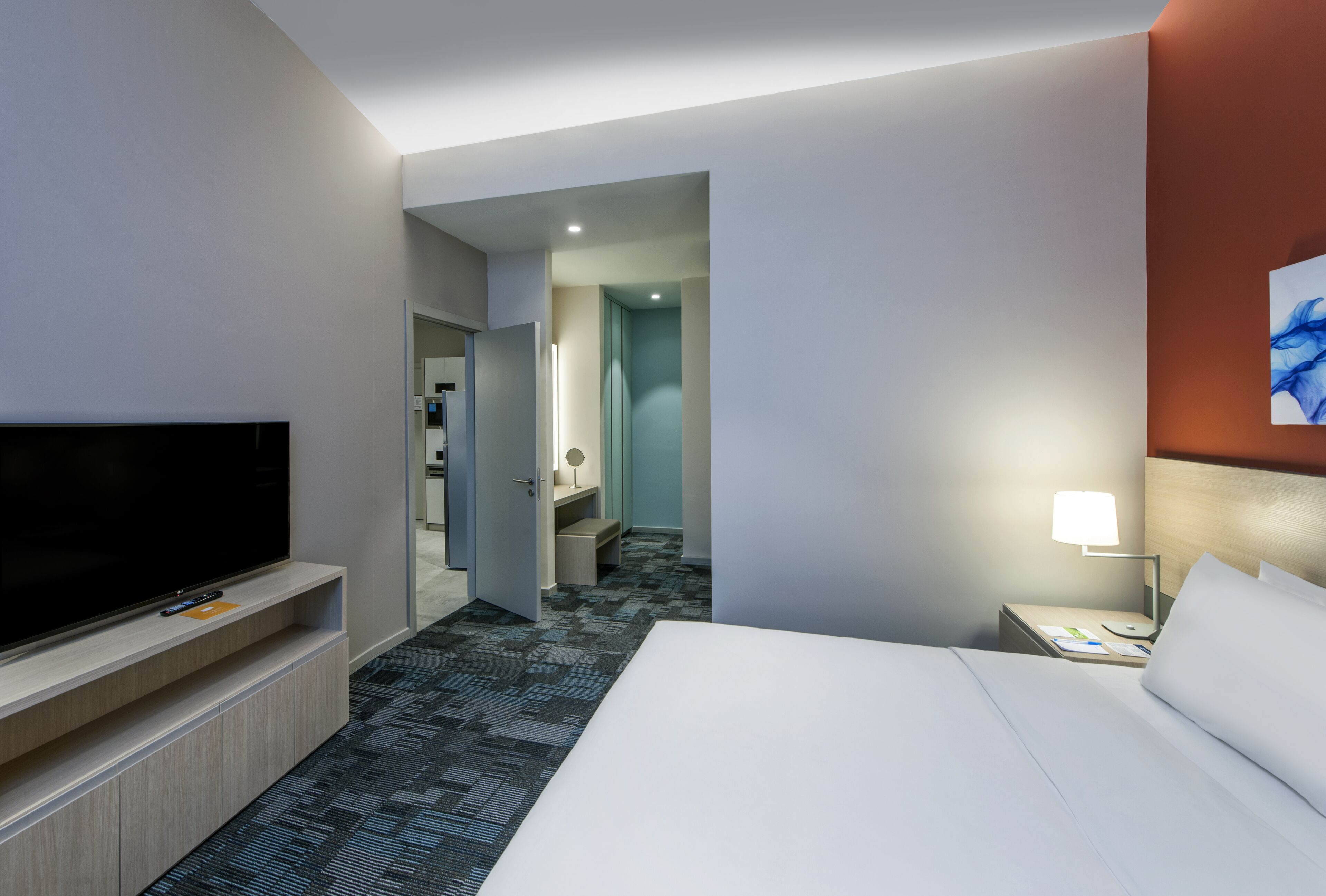A hotel room with a large bed, a flat-screen TV on a stand, a bedside table with a lamp, and a bathroom visible through an open door.