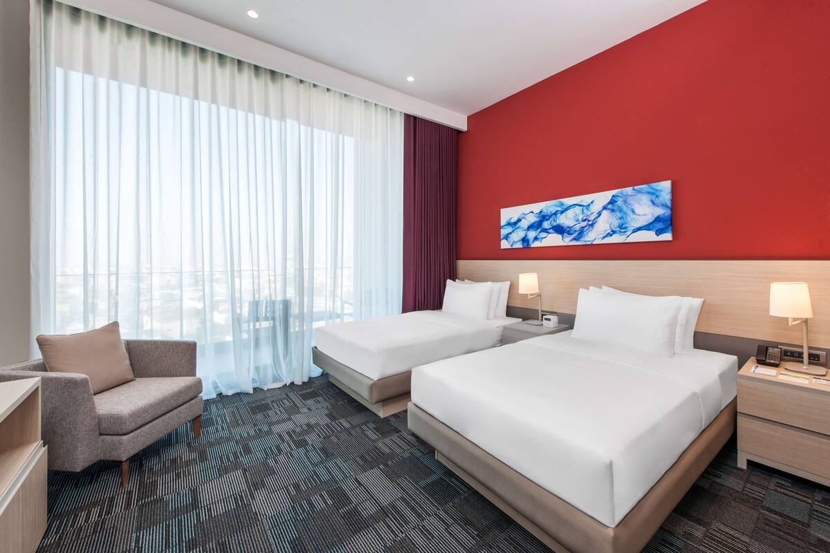 A hotel room with two beds, a sofa, a desk, and a large window with curtains.