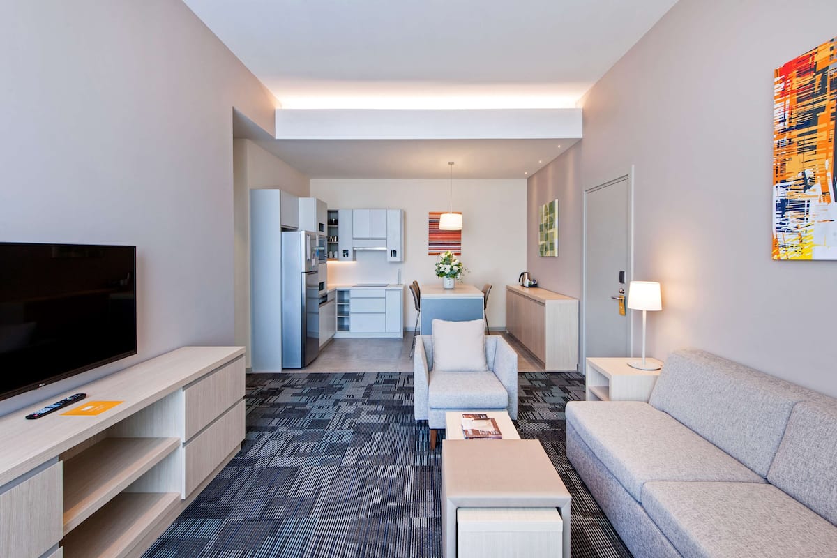 A modern hotel room with a flat-screen TV, a sofa, a dining area, and a kitchen.
